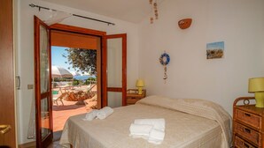 A bedroom with a bed, a bedside table with a lamp, a wall-mounted decoration, and a view of an outdoor area with umbrellas. - Sole 1 (Trinità d'Agultu e Vignola)