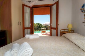 A hotel room with a bed, a wooden wardrobe, a small table with a lamp, and a view of a swimming pool and greenery outside.