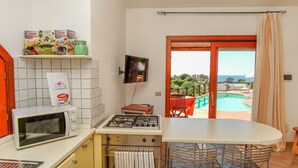 A kitchen with a microwave, stove, and a dining area with chairs. There is a TV mounted on the wall and a view of a swimming pool through an open door. - Sole 1 (Trinità d'Agultu e Vignola)