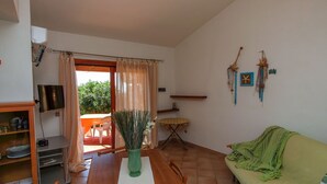 Un coin repas avec une table en bois, des chaises, un vase vert contenant des plantes séchées, un téléviseur et une porte-fenêtre coulissante donnant sur un espace extérieur.
