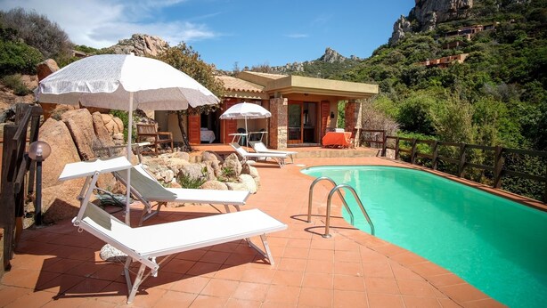 Un espace aménagé au bord de la piscine, avec des chaises longues et des parasols, une piscine et un paysage montagneux en arrière-plan.
