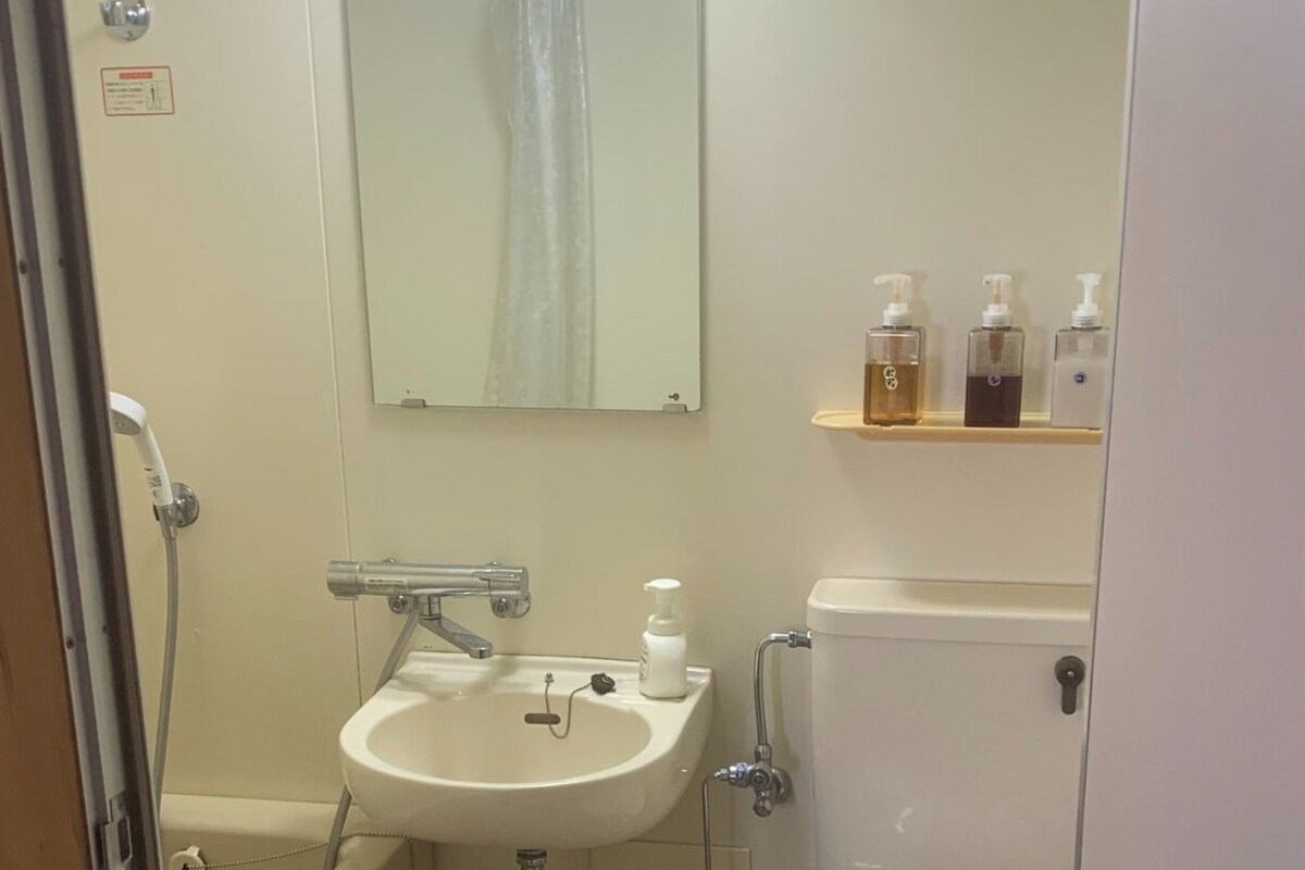 Une petite salle de bain avec un lavabo, des toilettes et une douche. On trouve des articles de toilette sur une étagère et des chaussons sur le sol.