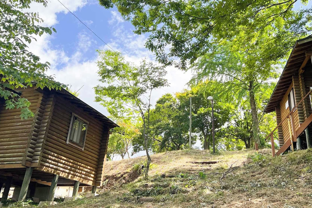 Two wooden cabins with log walls, surrounded by trees and a grassy hill.