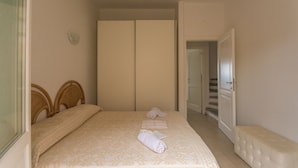A bedroom with a bed, two bedside tables, a bench, a wardrobe, and a door leading to a staircase. - Green 2 (Trinità d'Agultu e Vignola)