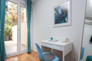 A room with a white desk, a chair, a framed abstract painting, and a view of a garden through a sliding door. - Sun Spalato Garden (Split)