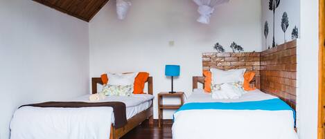 Una habitación con dos camas, cabeceros de madera, una mesita de noche con lámpara y un muro con motivos de hojas.