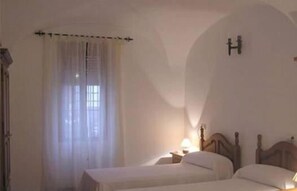 Un dormitorio con dos camas individuales, una mesita de noche, una lámpara, una ventana con cortinas traslúcidas y un aplique de pared.