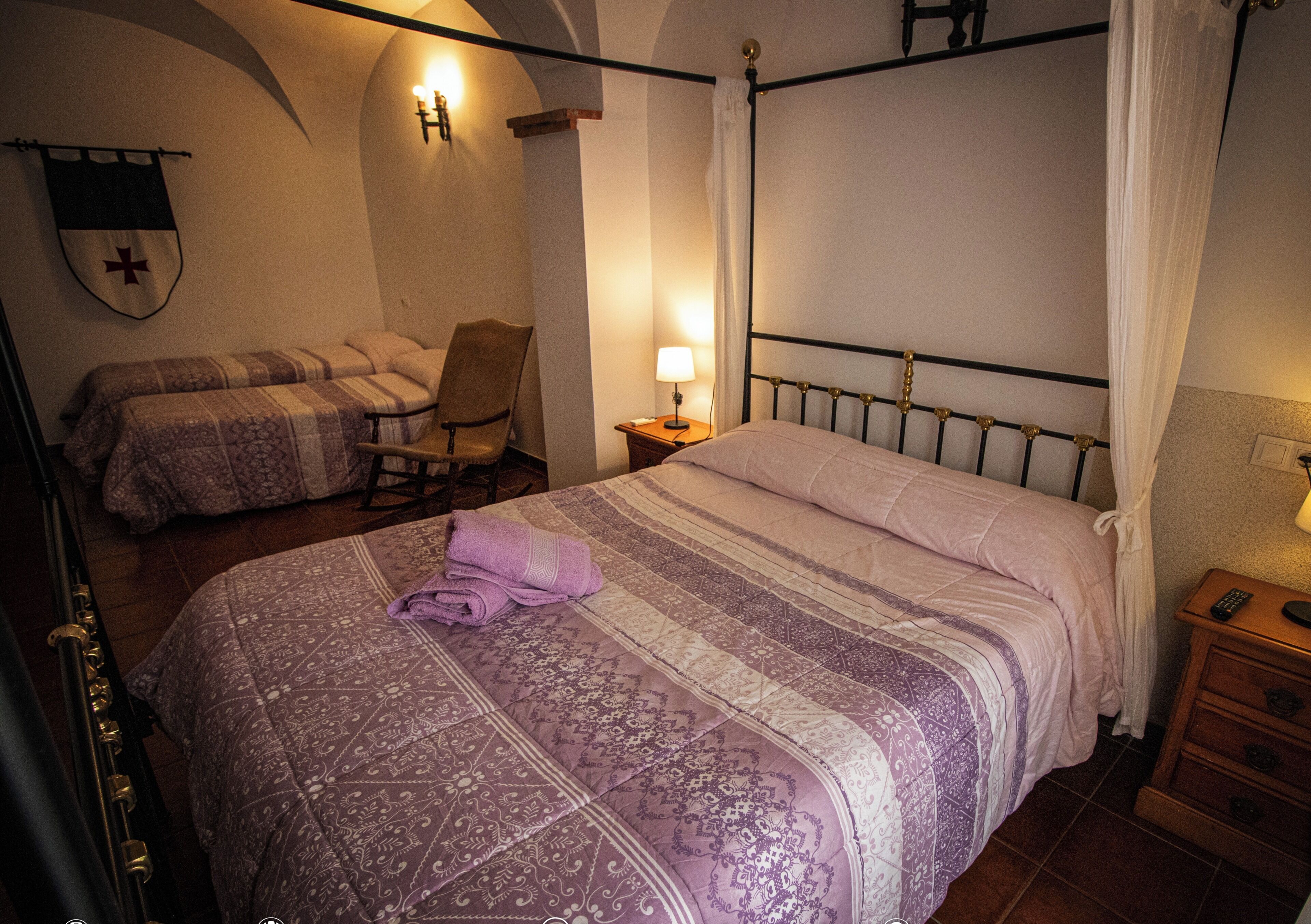 A bedroom with a four-poster bed, a chair, a bedside table with a lamp, and a wall-mounted cross.