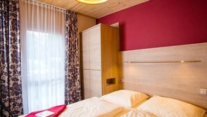 Ein Hotelzimmer mit Holzboden, einer roten Akzentwand, einem Bett mit weißen Leinentüchern, einem Kleiderschrank und einem Fenster mit durchsichtigen Vorhängen.