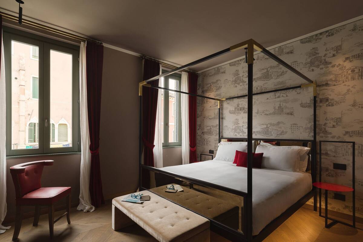 A four-poster bed with a wooden frame and white bedding, a red upholstered chair, a small wooden table, and a bench.