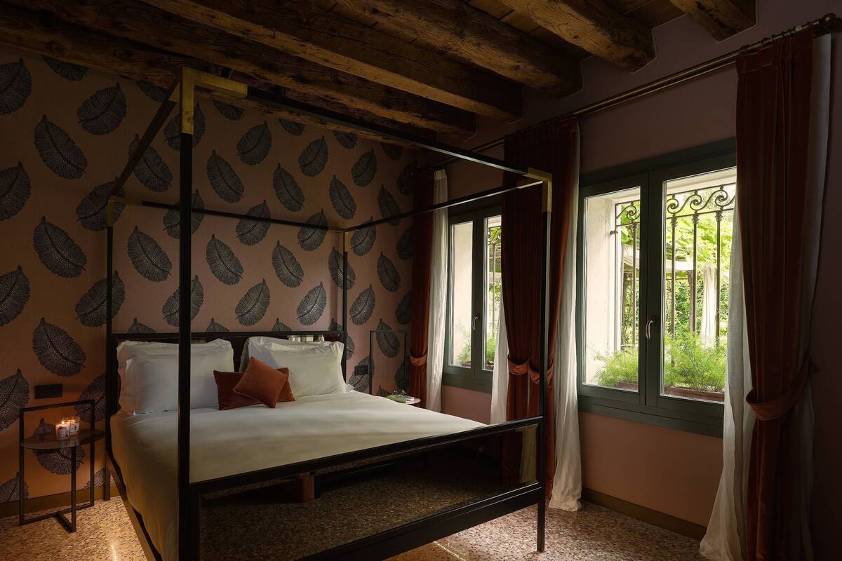 A four-poster bed with a leaf-patterned wallpaper, a window with curtains, and a view of greenery outside.
