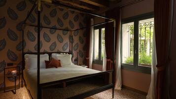 A four-poster bed with a leaf-patterned wallpaper, a window with curtains, and a view of greenery outside.