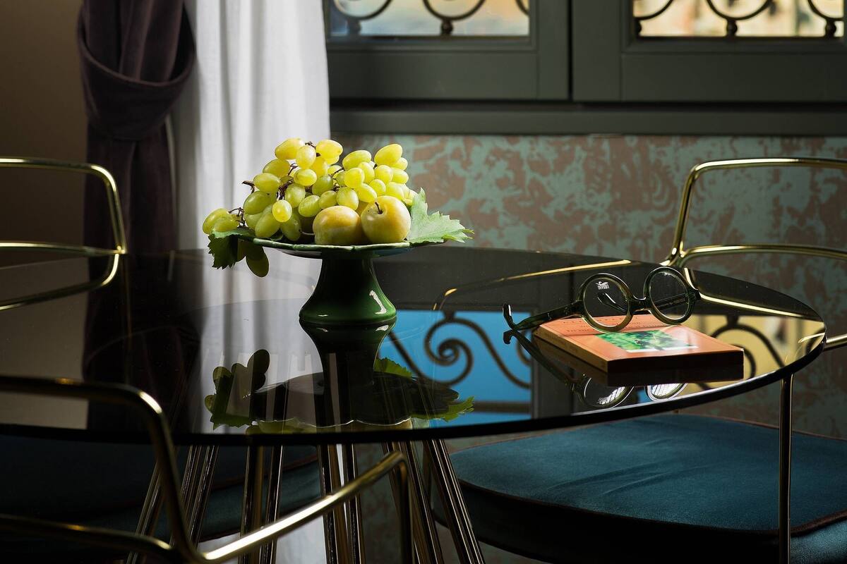 A table with a vase of yellow grapes and apples, a pair of glasses, and a book on a glass table near a window with a view of buildings.