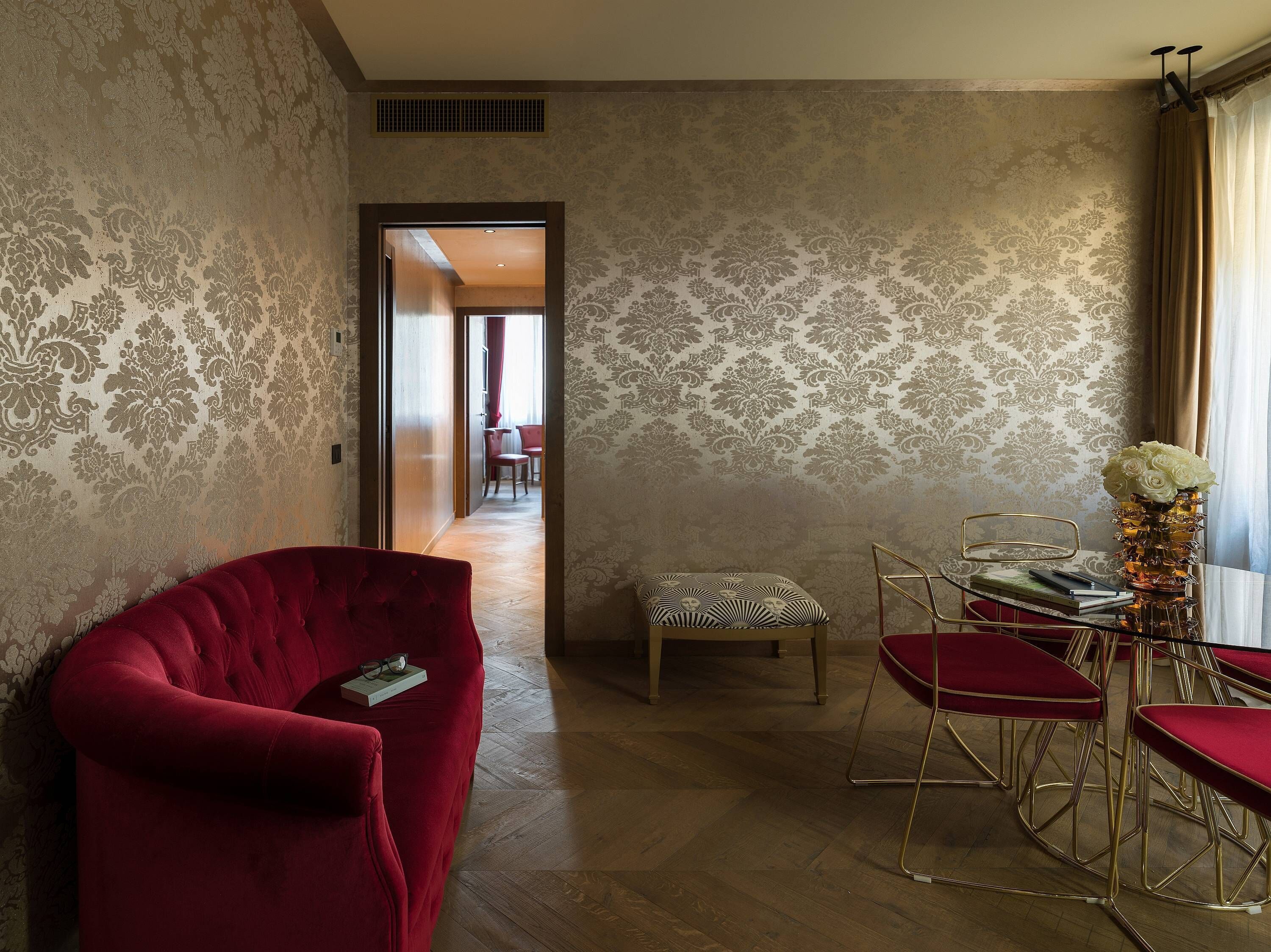Una habitación con papel tapiz estampado, un sillón tapizado en terciopelo rojo, una pequeña mesa con un jarrón de flores y vista a otra habitación con una silla roja y una mesa.