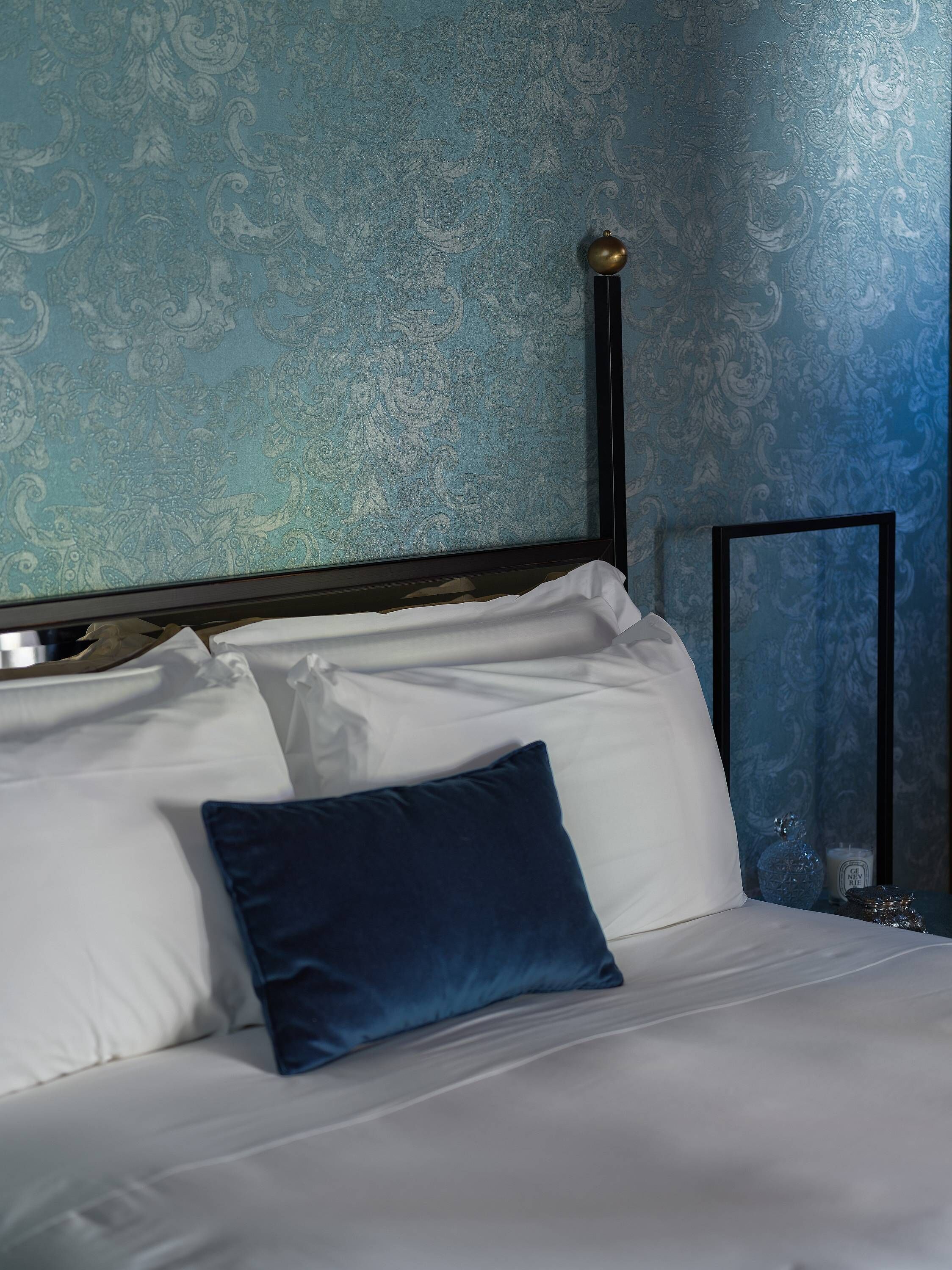 Una cama bien hecha con sábanas blancas y una almohada azul, cabecera de metal negro, y una pared con estampado paisley azul.