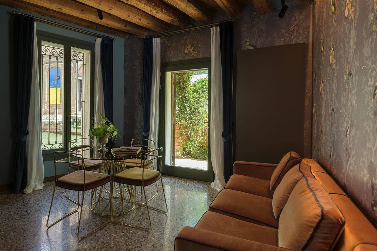 A living room with a wooden ceiling, a brown sofa, a dining table with chairs, and a view of greenery outside.