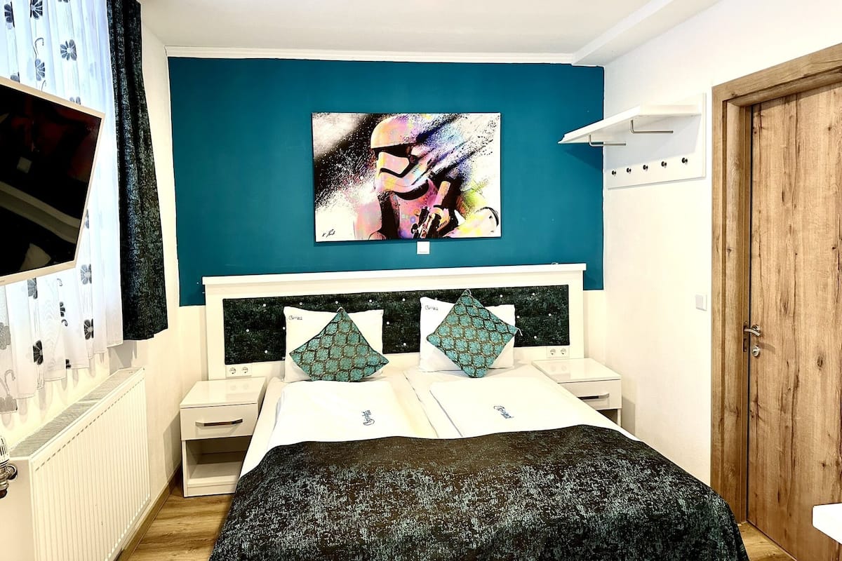 Un dormitorio con una pared color turquesa, una cama con colcha negra y verde, dos almohadas a juego, una mesita de noche blanca y un perchero de madera, además de un cuadro abstracto grande sobre la cama.