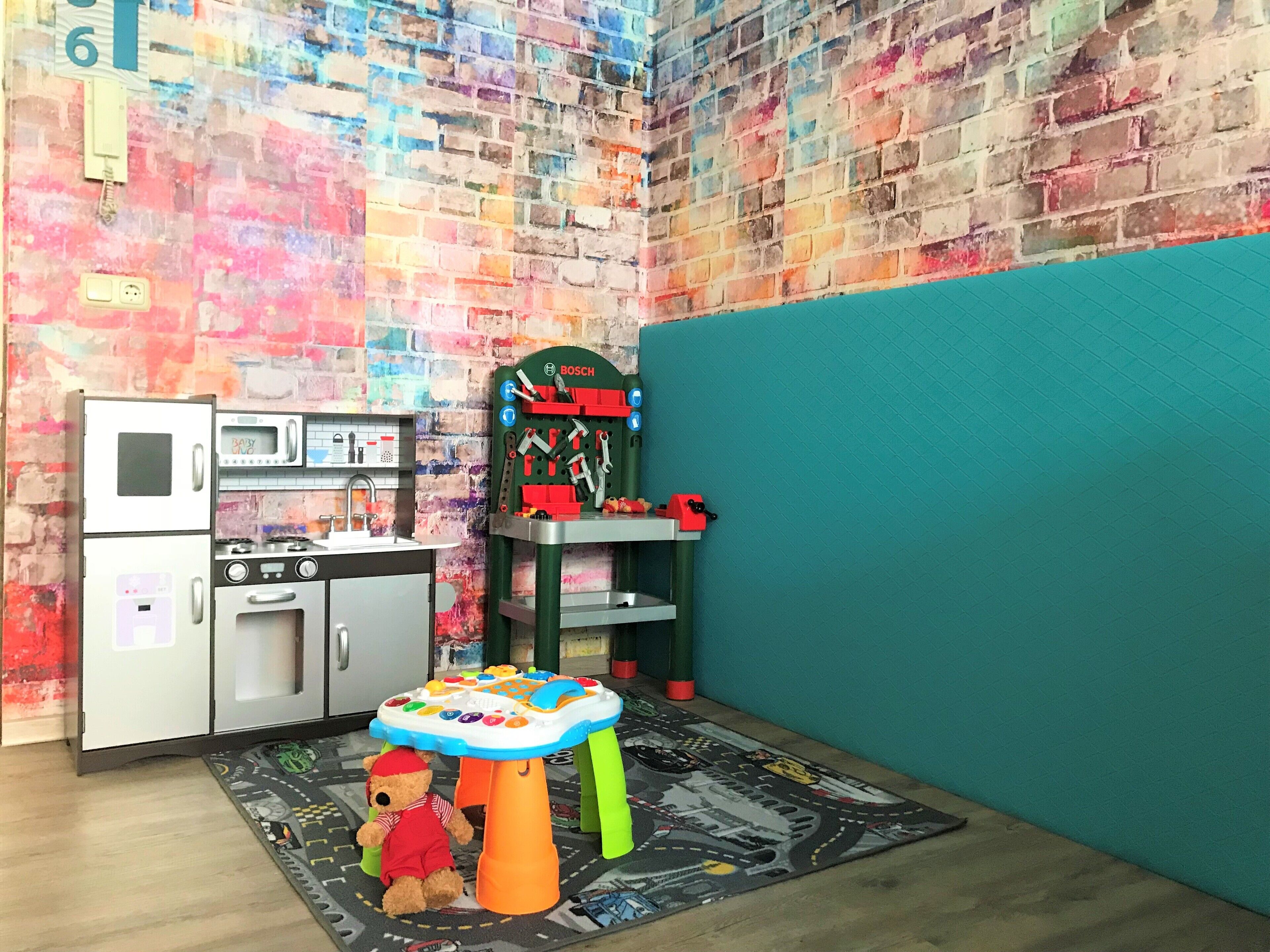 A play kitchen with a bear, a toy table, and a colorful wall.