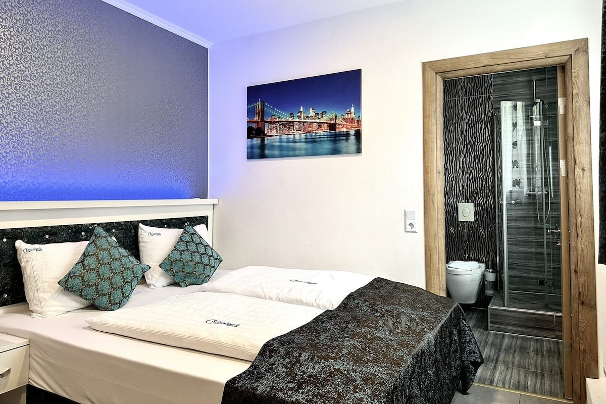 Una habitación de hotel con una cama, una mesita de noche, un cuadro en la pared de un paisaje urbano y una puerta de baño con ducha.