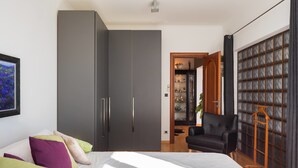 Un dormitorio moderno con una cama grande, un armario gris y una zona de estar con una silla negra y una mesita de madera.
