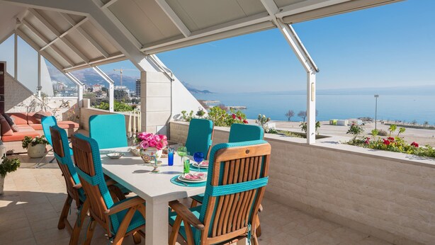 Un área de comedor al aire libre con vistas al mar, sillas de madera, una mesa dispuesta con platos y un porche cubierto.