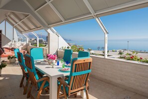 Un área de comedor al aire libre con vistas al mar, sillas de madera, una mesa dispuesta con platos y un porche cubierto.