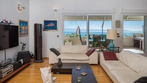 Una sala de estar moderna con un sofá, una mesa de centro y un televisor de pantalla plana. Se ve una ventana grande con vista al mar.