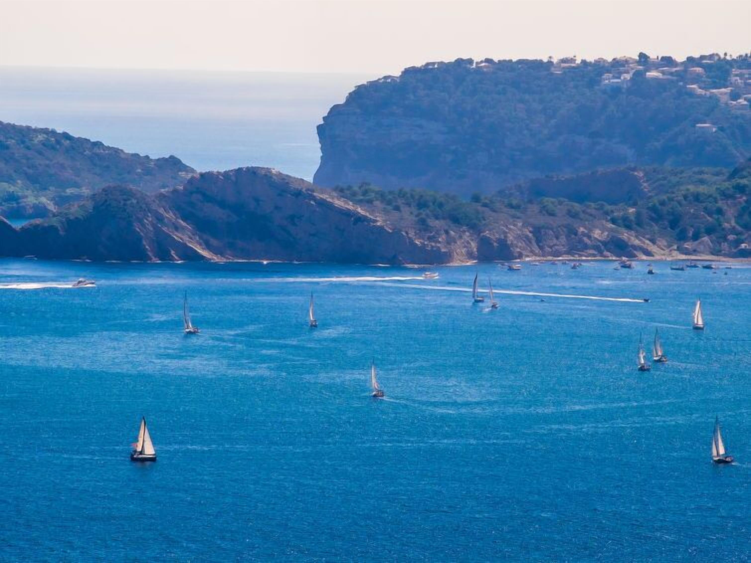 A coastal scene with multiple sailboats on the water, cliffs in the background, and a clear sky.