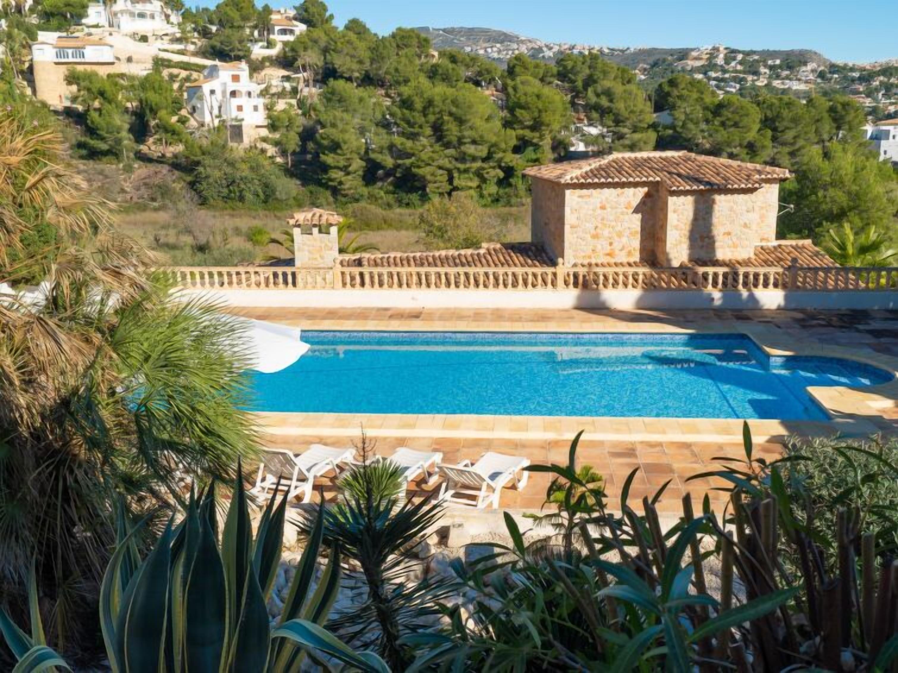 A pool area with a stone building, surrounded by greenery and overlooking a hillside.