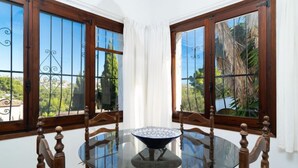 A dining area with a round glass table and chairs, large windows with metal grilles, and a view of greenery outside. - Sunshine by Interhome (Moraira)