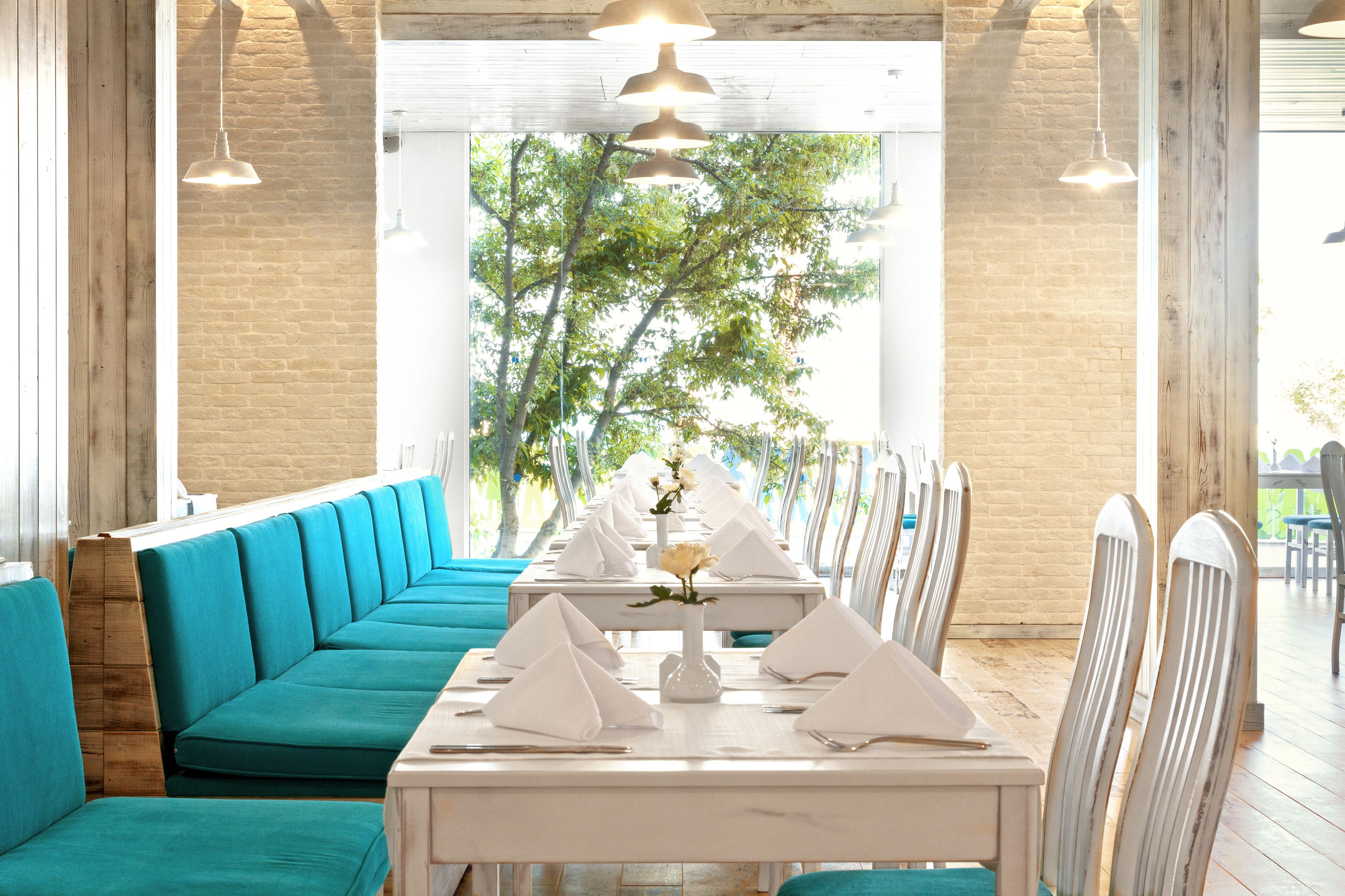 Un restaurante con una mesa larga dispuesta para una comida, asientos con cojines color turquesa y vistas a árboles a través de un amplio ventanal.