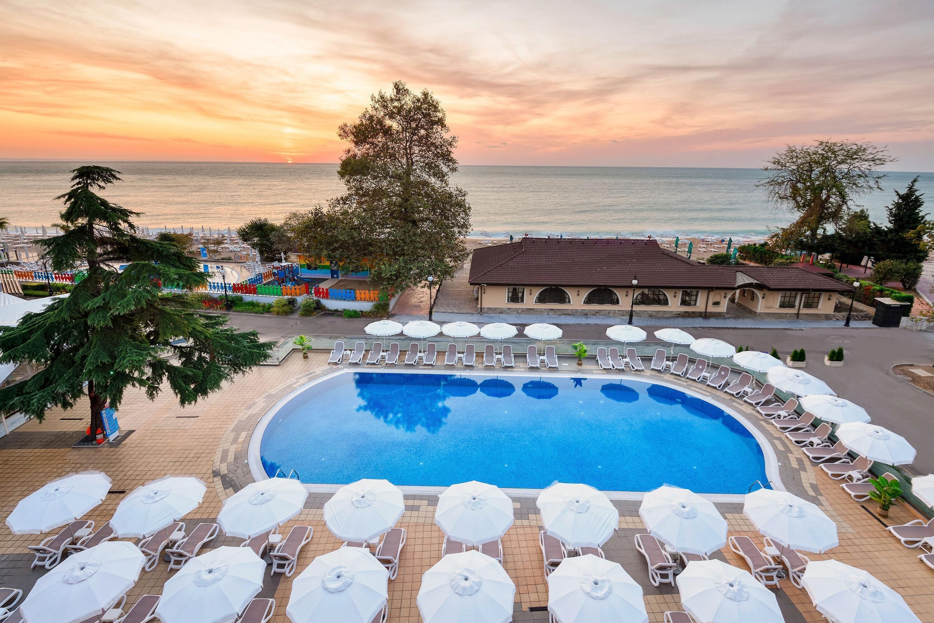 A poolside area with sun loungers and umbrellas, a clear view of the sea, and a building with a tiled roof.