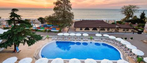 A poolside area with sun loungers and umbrellas, a clear view of the sea, and a building with a tiled roof.