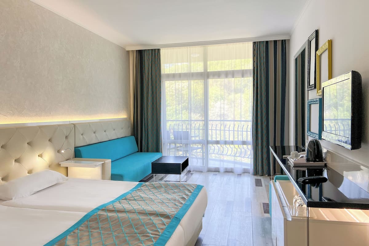 A hotel room with a large bed, a sofa, a desk with a chair, a TV, and a balcony with a view.