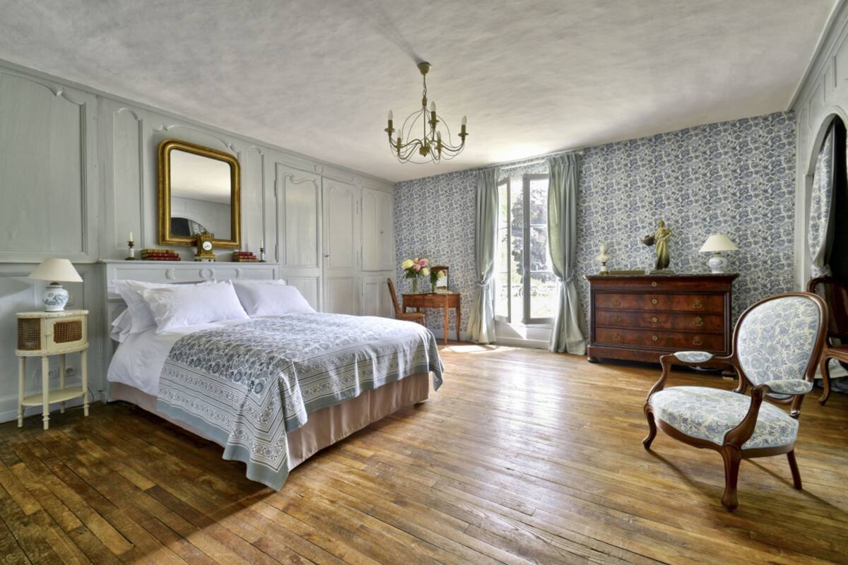 Ein Schlafzimmer mit einem großen Bett, Holzboden, einem Schminktisch, einem Sessel und einer Deckenleuchte.