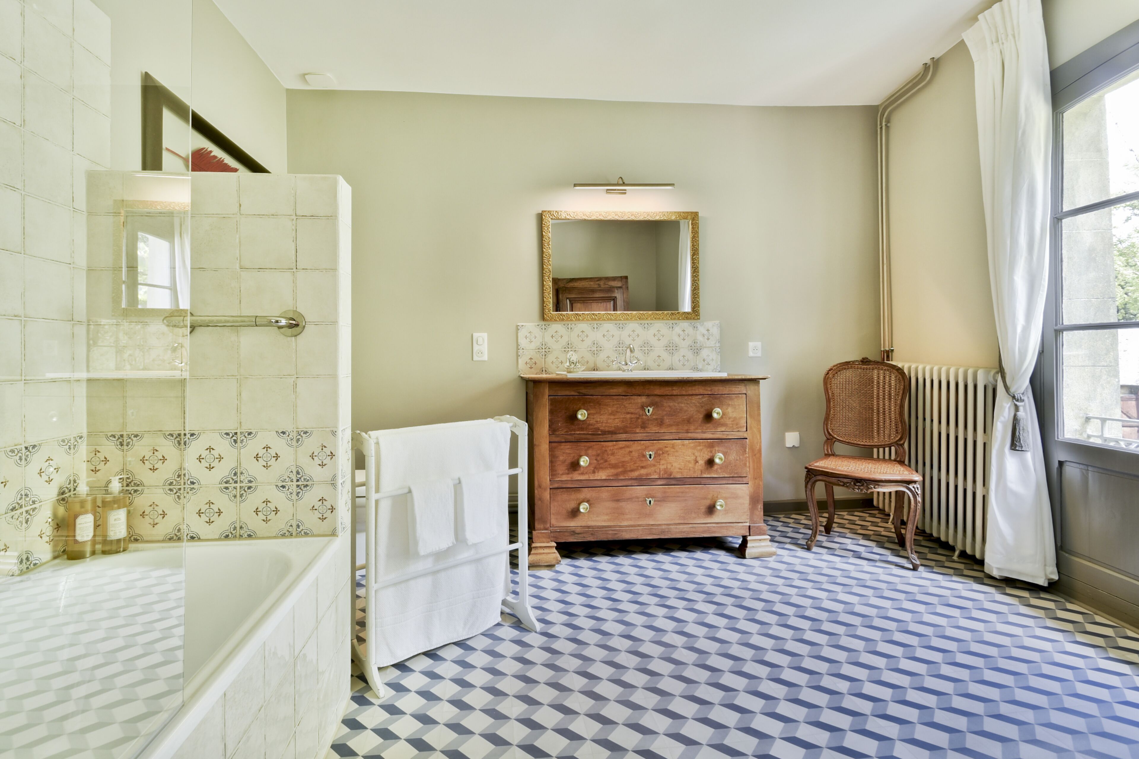Een badkamer met een bad, een houten dressoir, een rieten stoel en vloertegels met een patroon.