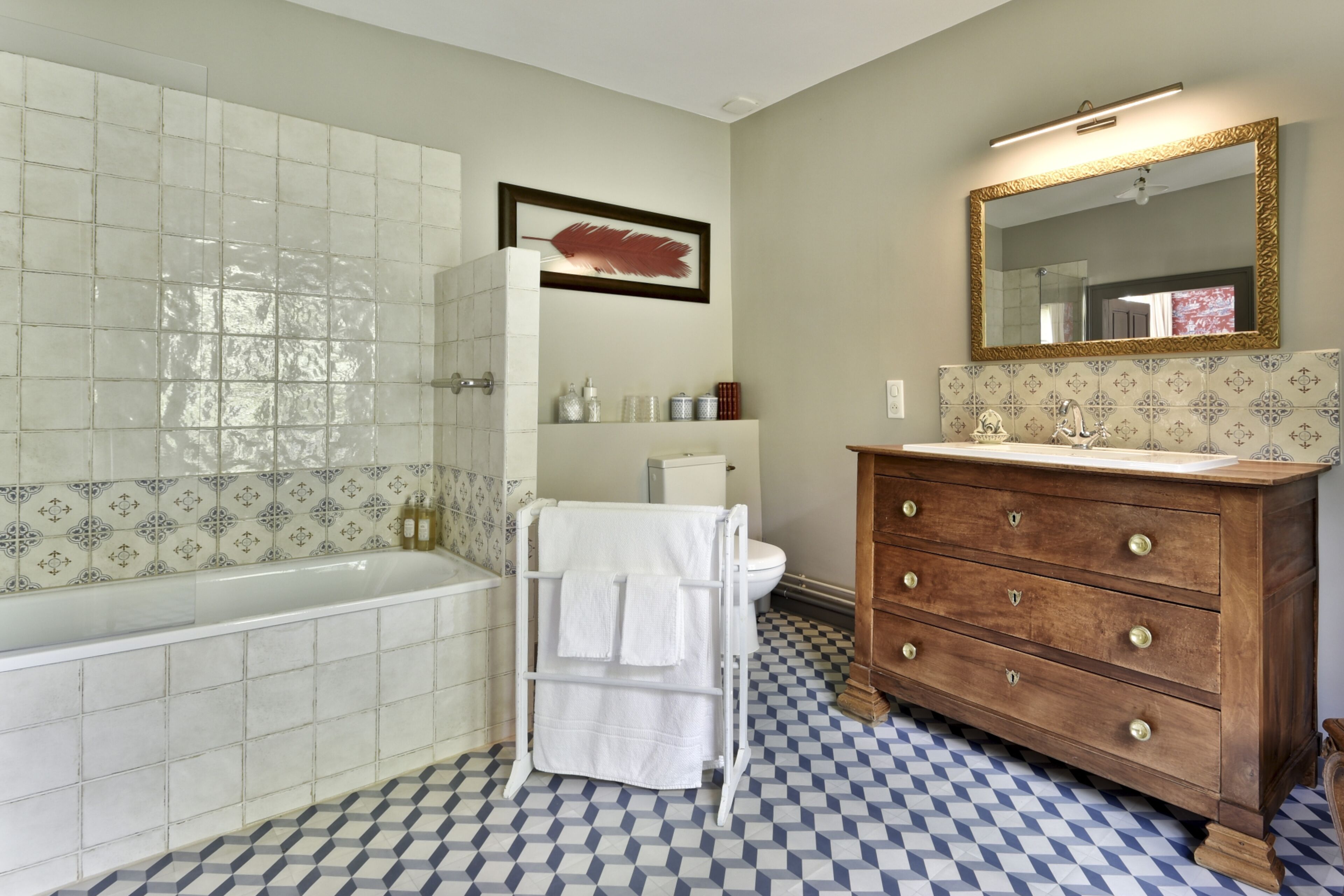 A bathroom with a checkered floor, a white bathtub, a wooden vanity with a mirror, and a framed artwork on the wall.