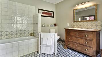 A bathroom with a checkered floor, a white bathtub, a wooden vanity with a mirror, and a framed artwork on the wall.