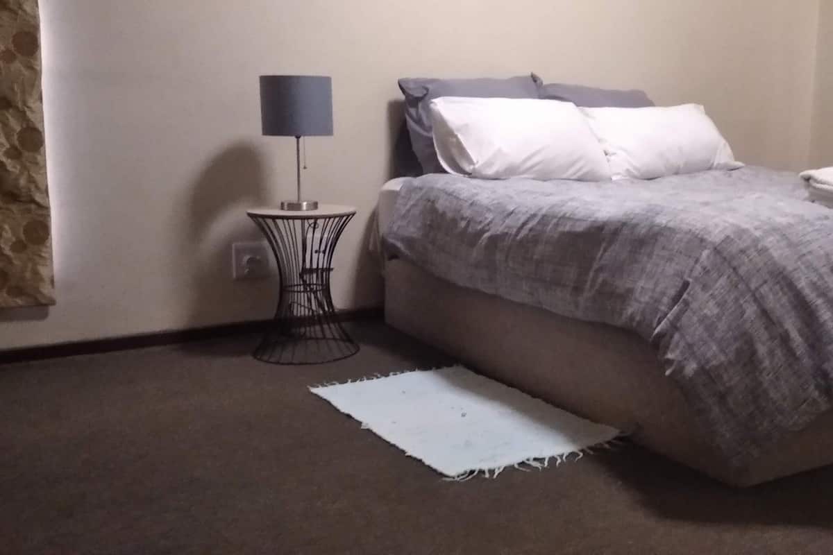Un dormitorio con cama, una mesita de noche con lámpara, una alfombra blanca y una ventana con cortinas.