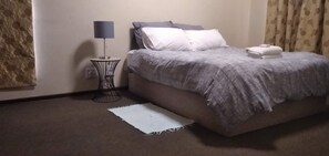 Ein Schlafzimmer mit einem Bett, einem Nachttisch mit Lampe, einem weißen Teppich und einem Fenster mit Vorhängen.