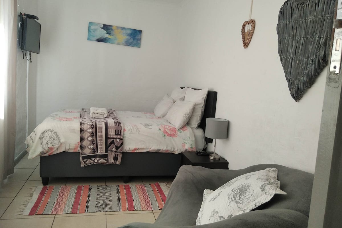 Un dormitorio con cama, mesita de noche, lámpara, sofá, televisor montado en la pared y un cuadro.