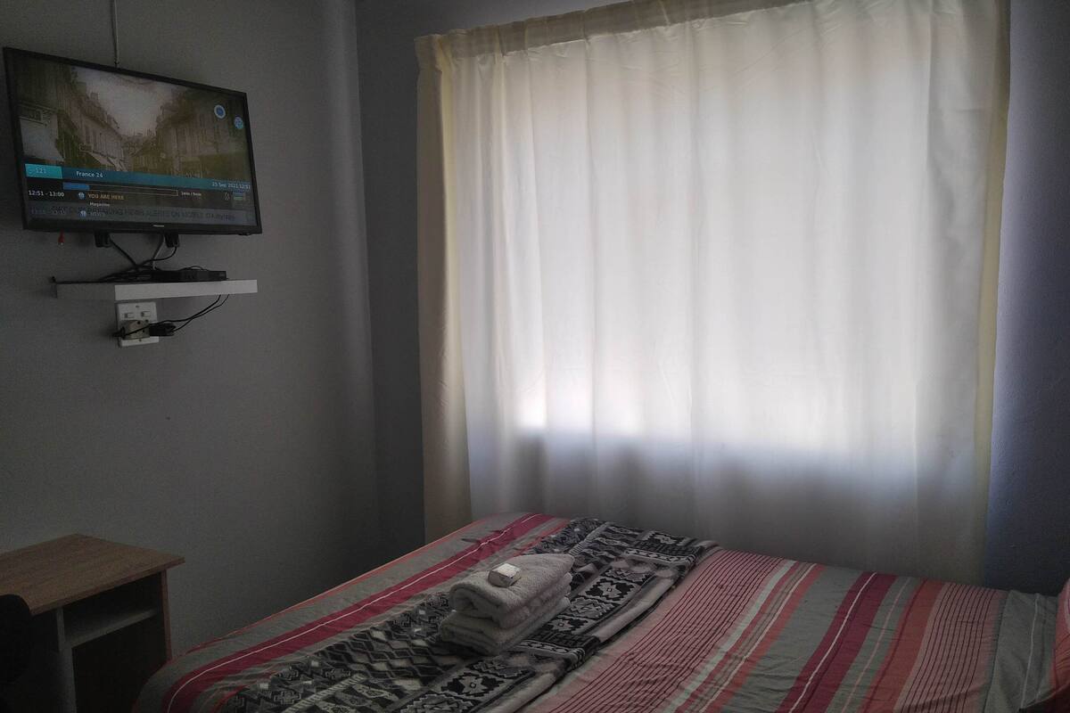 Una habitación de hotel con cama, televisor montado en la pared y una ventana con cortinas.