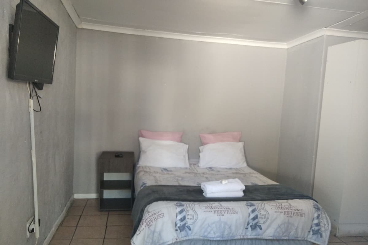 Un dormitorio con cama, almohadas, una mesita de noche, un televisor montado en la pared y una puerta.