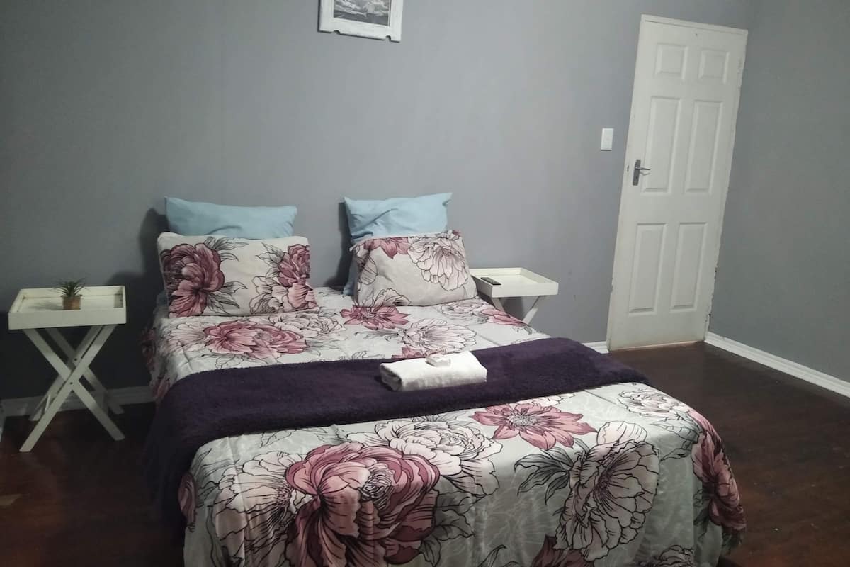 Un dormitorio con cubrecama floral, dos almohadas, una mesita de noche con una planta y una puerta.