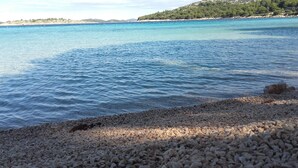 Una playa de guijarros con agua azul cristalina y una colina distante cubierta de árboles.