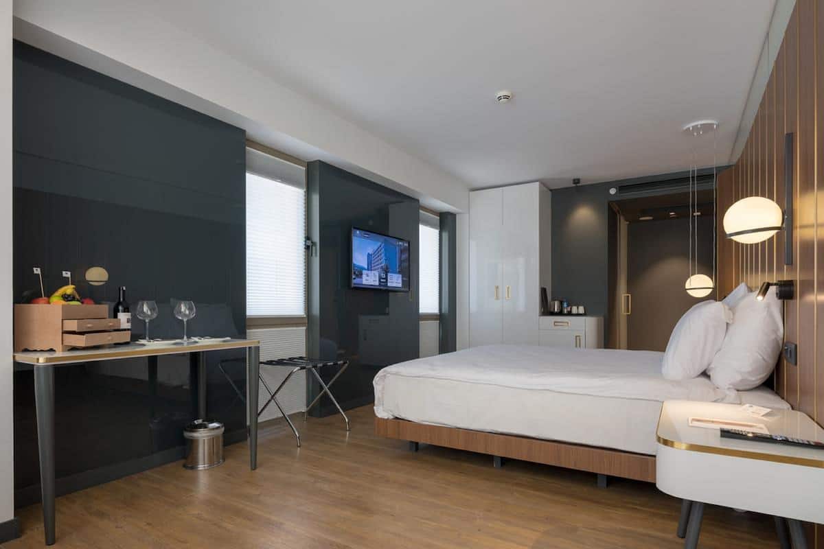 A modern hotel room with a bed, bedside table, TV, and a desk with a chair.