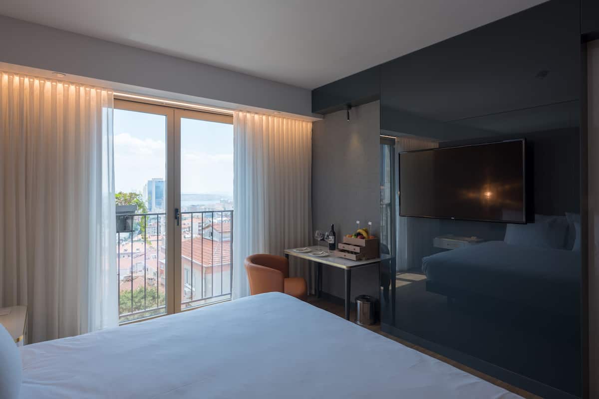A modern hotel room with a large bed, a desk with a chair, a TV mounted on the wall, and a balcony with curtains.