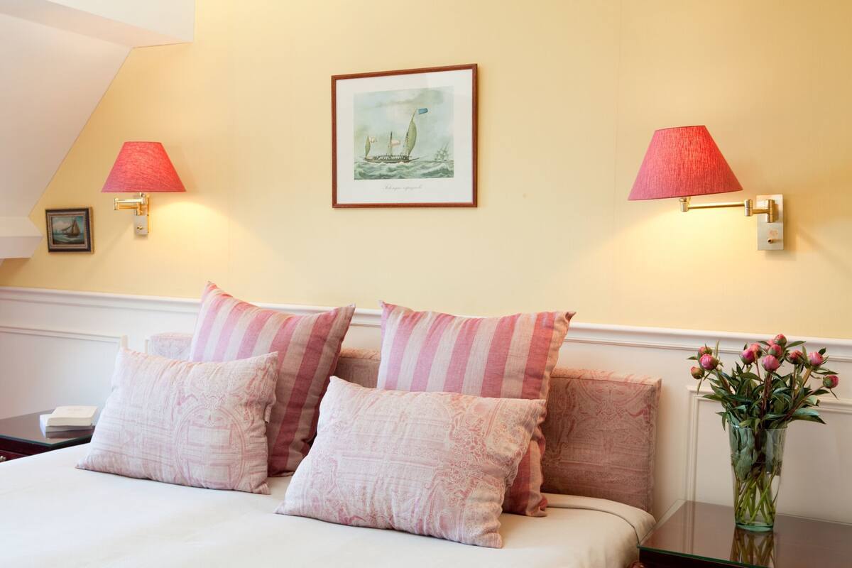 A neatly made bed with pink and white striped pillows, a framed seascape painting above, and a vase with pink flowers on a nightstand.