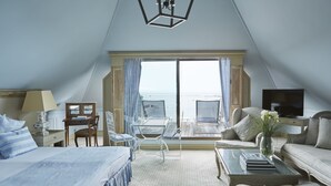 Habitación de hotel con cama, escritorio, silla, sofá, mesa de centro y televisor. La habitación tiene un ventanal con vistas al mar.