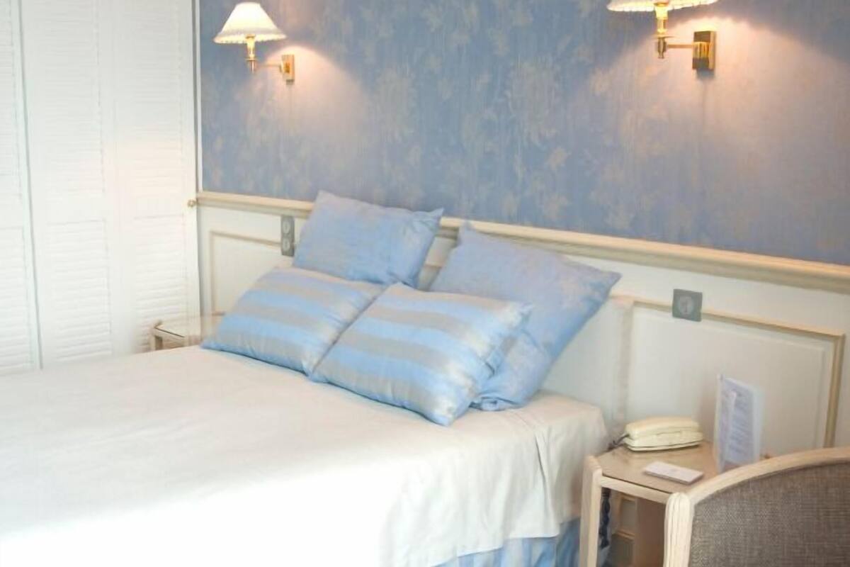 A hotel room with a bed, two wall-mounted lamps, a bedside table with a lamp, and a chair.